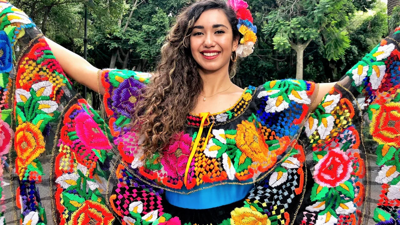 Smiling woman wearing a richly embroidered, multicolored folk dress and floral hair adornment, set against a leafy backdrop.