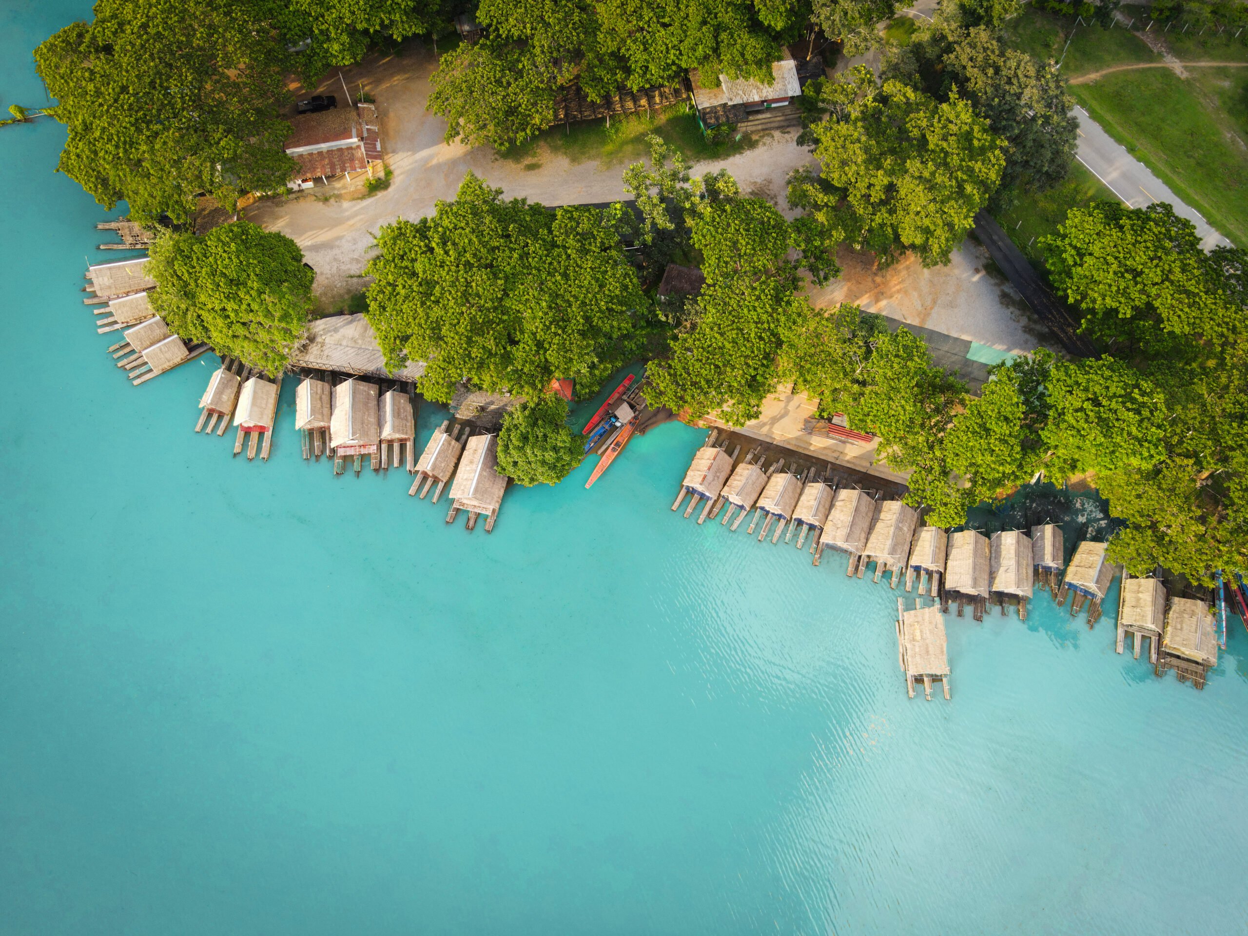An aerial view of turquoise blue water lined with bamboo raft houses along a lush green forested shoreline. The scene captures wooden huts elevated on stilts over the water, with a few traditional boats docked nearby. The pristine blue water contrasts beautifully with the surrounding greenery, creating a serene and picturesque landscape, ideal for eco-tourism and nature retreats.