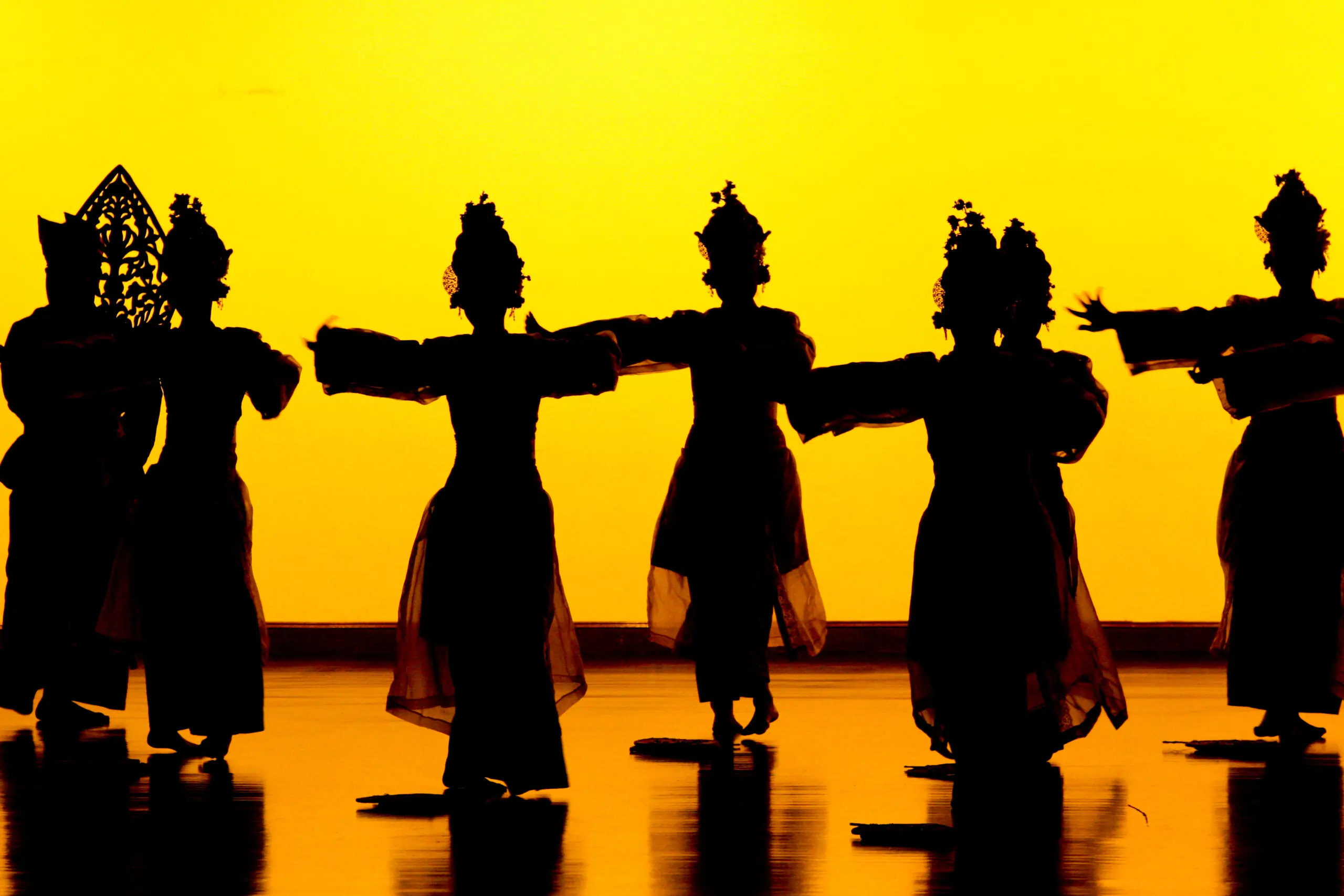 Silhouette of dancers performing traditional dances of Andaman and Nicobar Islands against a vibrant yellow-orange background.